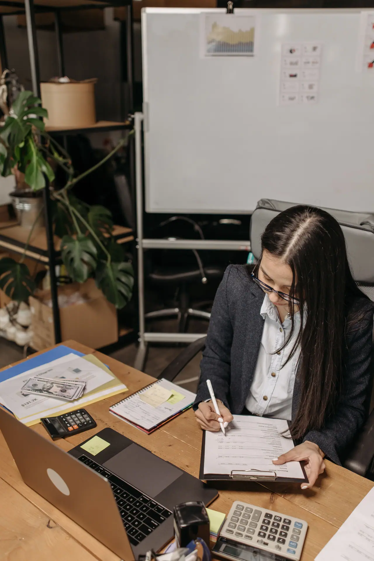 Hands completing a balance sheet on a clipboard at a wooden desk, with a laptop, a calculator and a notebook alongside.